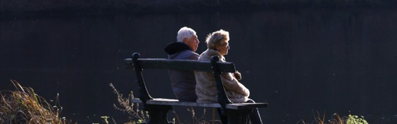 An older couple sitting together on a park bench in the sunshine, illustrating Medicare plan review in retirement.