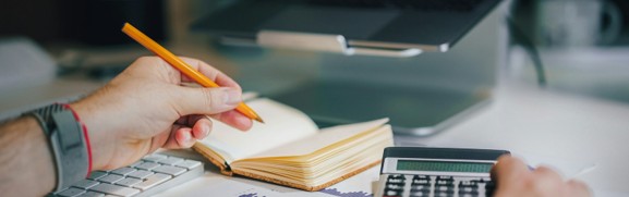 Hands using a calculator and notebook beside printed financial charts to compare self-employed health coverage.